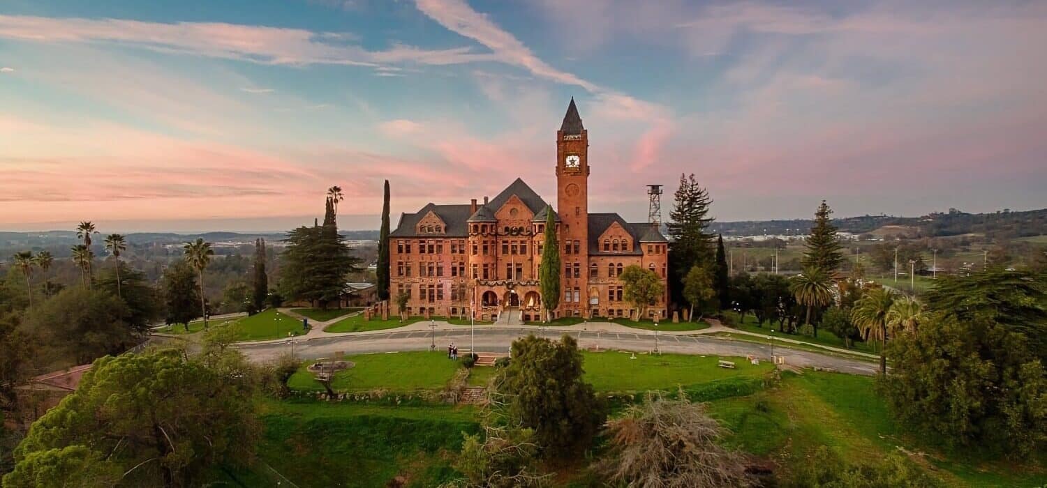 a red brick castle with a tall clock tower amidst green grass, bushes and trees, and a blue and pink sky