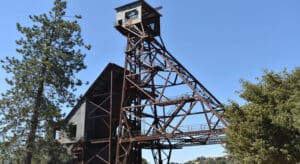 a mine shaft and tower next with a pine tree and green bushes on either side, and blue skies above